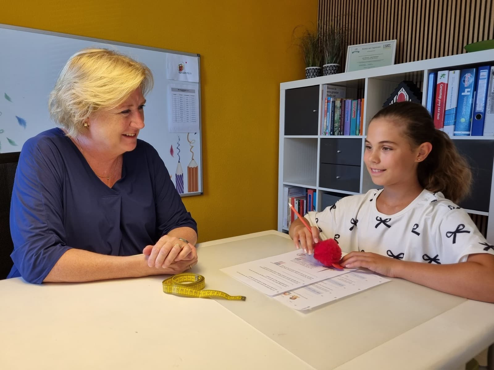 Ina zit aan tafel naast een meisje dat met een potlood aan een rekenopdracht werkt.