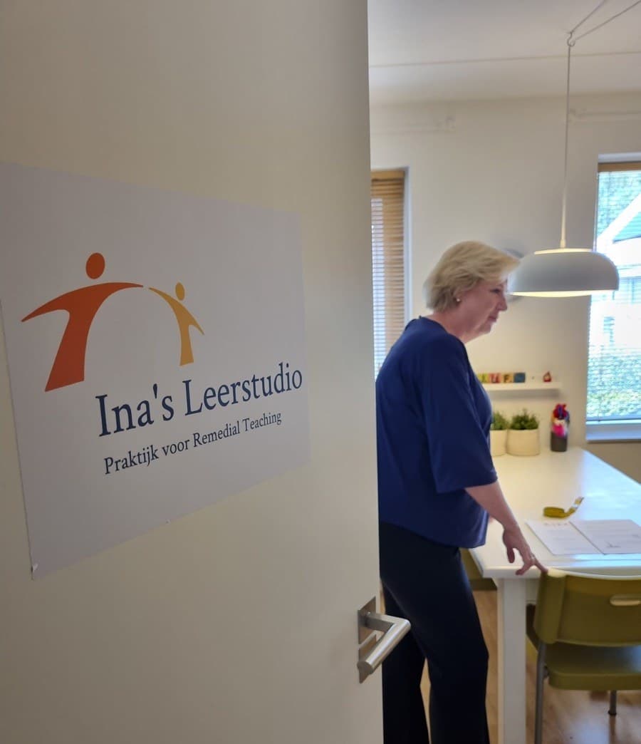 Ina staat bij de deur van de leerstudio, met het bordje van Ina's Leerstudio ernaast.